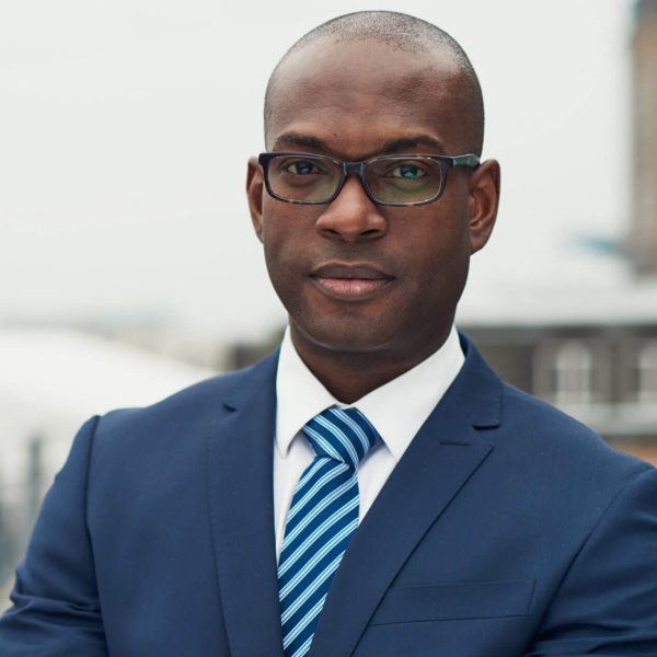 Confident black business man in a stylish suit standing with folded arms on a rooftop of n office block looking at the camera with a serious expression